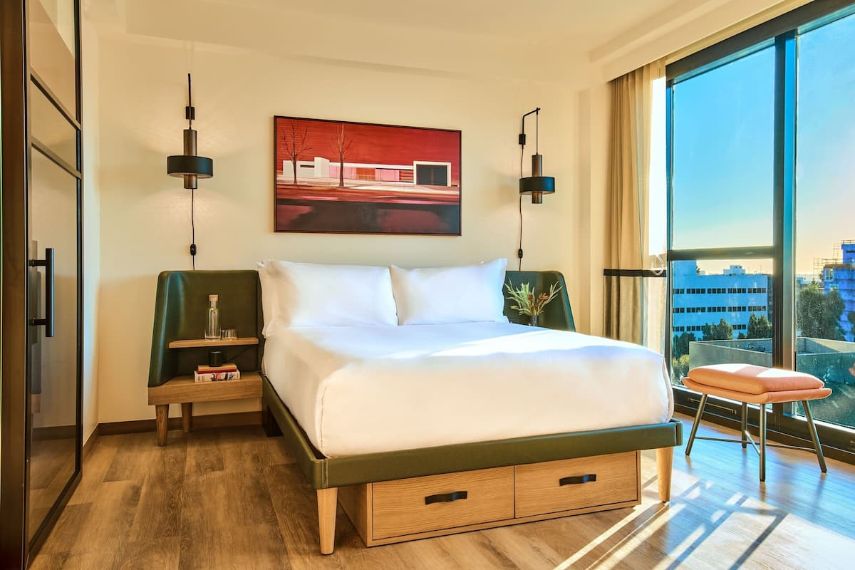 A modern hotel room with a large bed, bedside tables, a nightstand, a window with a city view, and a painting on the wall.