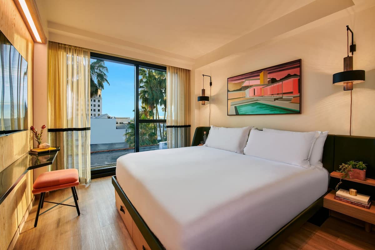 A hotel room with a large bed, a desk with a chair, a window with curtains, a painting on the wall, and a nightstand with a lamp.