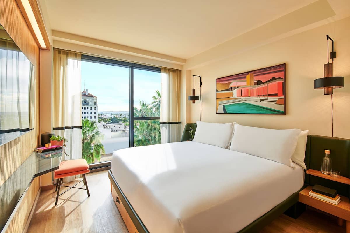 A hotel room with a large bed, a desk with a chair, a painting on the wall, and a view of the city through the window.