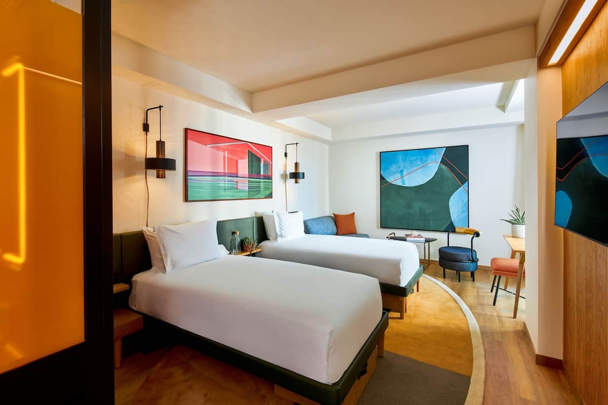 A hotel room with two beds, a desk, a chair, and artwork on the walls.