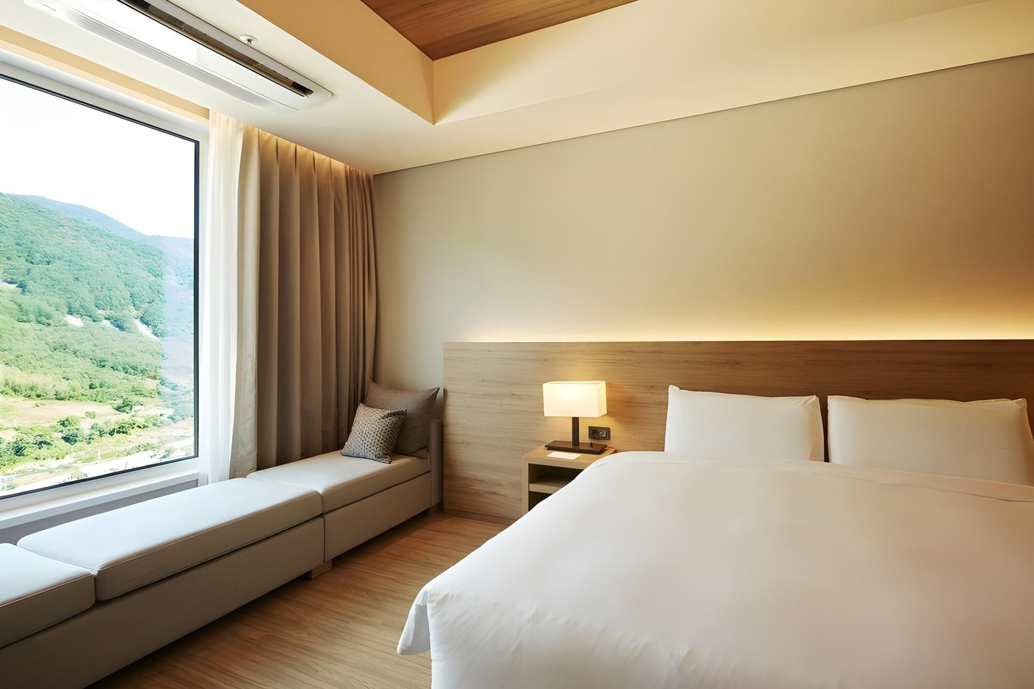 Quarto de hotel com uma cama grande, um assento junto à janela com cortinas, uma mesa de cabeceira com abajur e vista para uma paisagem verde.