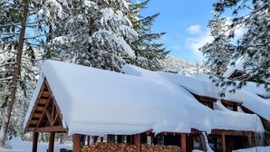 Eine Holzhütte mit schneebedecktem Dach, Holzfassade und einem Holzstapel davor.