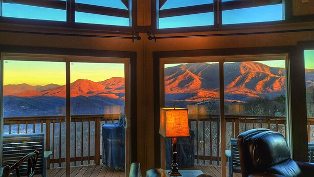 A room with large windows offering a mountain view, a wooden deck, and a table with chairs.