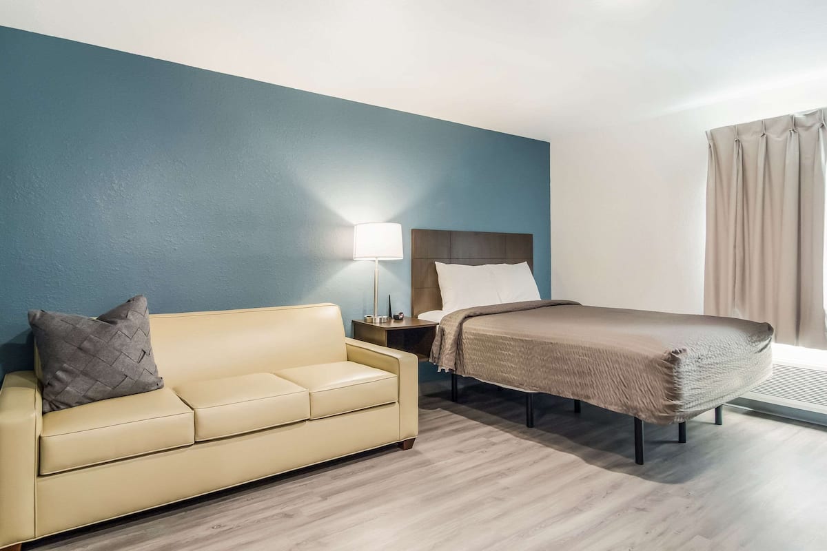 A hotel room with a beige sofa, a bed with a brown blanket, a nightstand with a lamp, and a window with curtains.