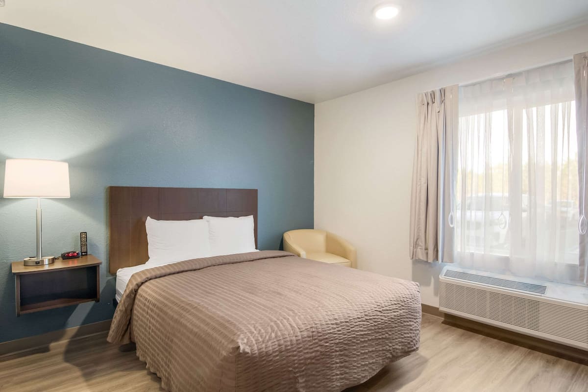 A hotel room with a bed, a nightstand with a lamp, a chair, and a window with curtains.
