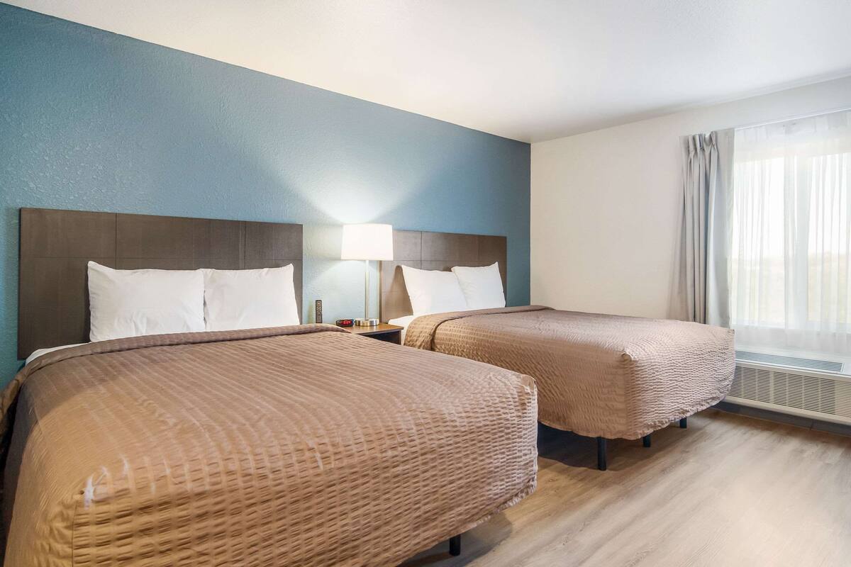 A hotel room with two beds, a nightstand with a lamp, and a large window with curtains.