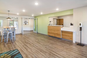 A reception area with a wooden reception desk, a counter, and a printer. There is a seating area with chairs and a table. The room has wooden flooring and a green accent wall. - WoodSpring Suites Jacksonville Baymeadows (Jacksonville)