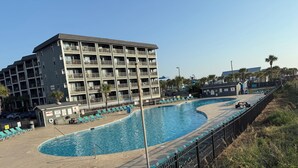A swimming pool with lounge chairs, a building with multiple balconies, and a fenced area with palm trees. - OCEANFRONT 1st floor unit! 150 steps away from the sand AND multiple pools!! (Myrtle Beach)
