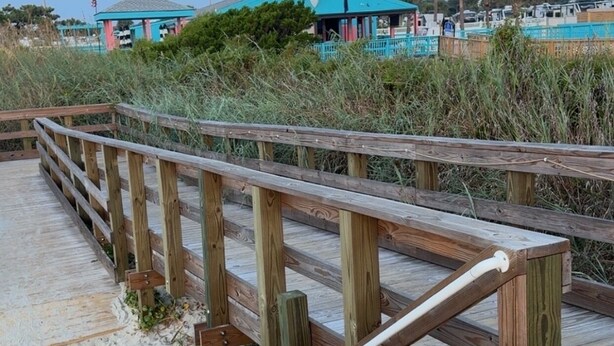 A wooden boardwalk leading to a building with a blue roof, surrounded by sand and vegetation. - OCEANFRONT 1st floor unit! 150 steps away from the sand AND multiple pools!! (Myrtle Beach)