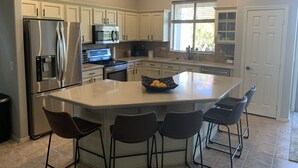 A modern kitchen with white cabinets, stainless steel appliances, a large island with bar stools, and a clock on the wall. - Four bedroom home on golf course. (Mesa)