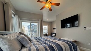 Un dormitorio con una cama grande, un ventilador de techo, un televisor montado en la pared y vistas al exterior.