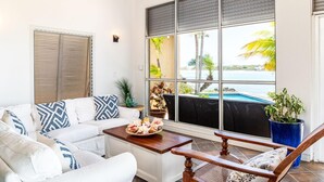 Ein sonnendurchfluteter Raum mit einem weißen Sofa, einem Holztisch und Blick auf einen Pool und Palmen.