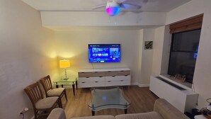A living room with a sofa, a glass coffee table, a wooden chair, a TV mounted on the wall, and a ceiling fan.