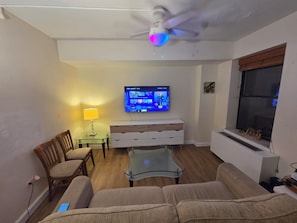 A living room with a sofa, a glass coffee table, a wooden chair, a TV mounted on the wall, and a ceiling fan.