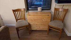 A small wooden table with two chairs, a window with a view of buildings, and a white container on the windowsill.