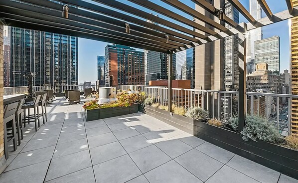 A rooftop terrace with a pergola, seating area, and a view of surrounding high-rise buildings.