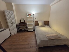 A bedroom with a bed, a desk with a chair, a wardrobe, and a shelf with a lamp.