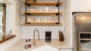 Una cocina con salpicadero de azulejos blancos, estantes de madera, un refrigerador de acero inoxidable, una cafetera y un lavamanos.