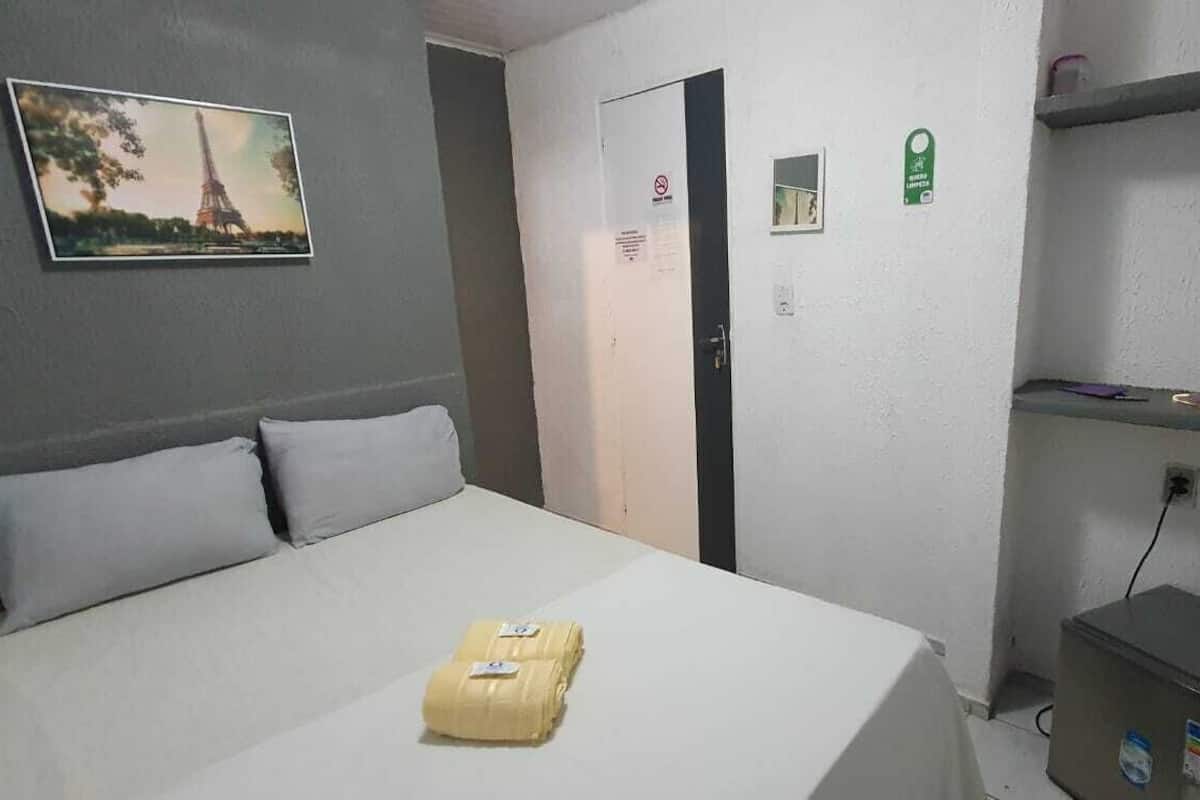 A hotel room with a bed, a small desk, a shelf with items, a door, and a picture on the wall.