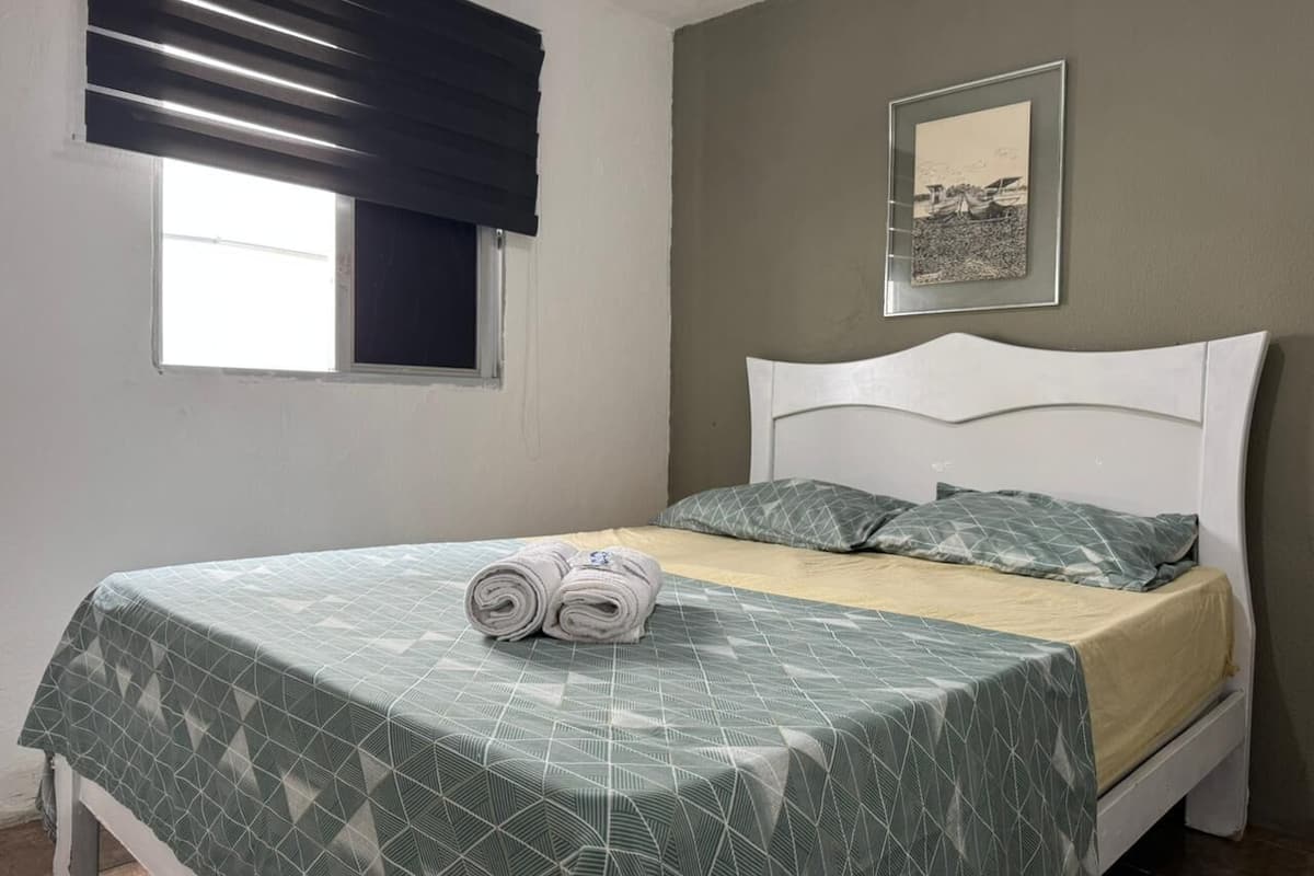 A single bed with a geometric patterned bedspread, a white headboard, and two pillows. There is a framed picture on the wall above the bed.