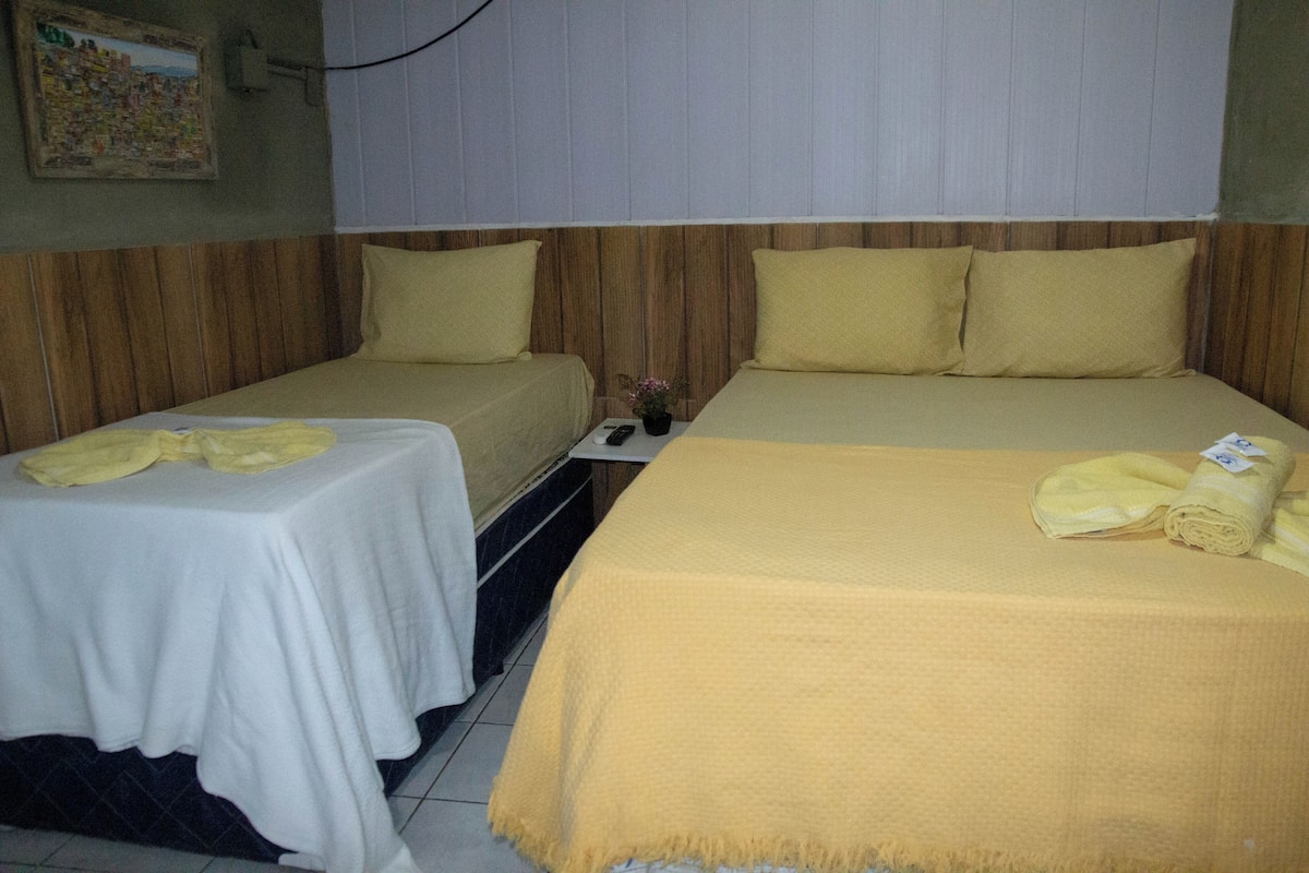 A hotel room with two single beds, a large bed, and a small table with a remote control.