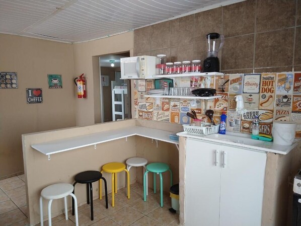 Una cocina con encimera blanca, una licuadora y una cafetera. Hay tres taburetes y un extintor en la pared.