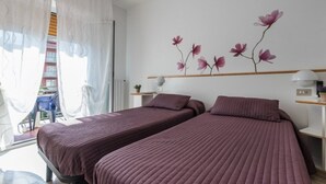 Una habitación con dos camas, un ventanal con cortinas traslúcidas, una pared decorada con un mural floral y una mesita de noche con una lámpara.