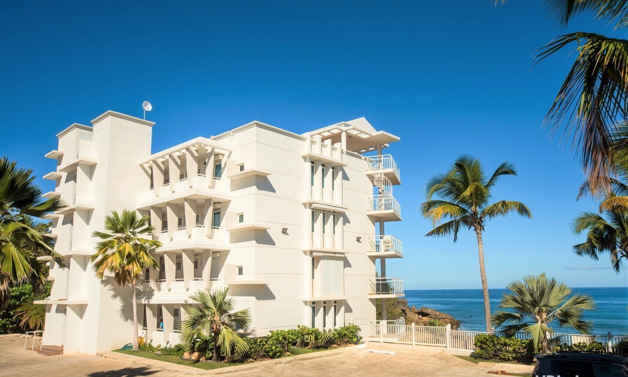 A multi-story hotel with balconies, located near a beach with palm trees.
