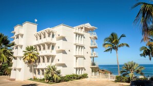 Un hôtel à plusieurs étages, doté de balcons, situé près d’une plage bordée de palmiers.