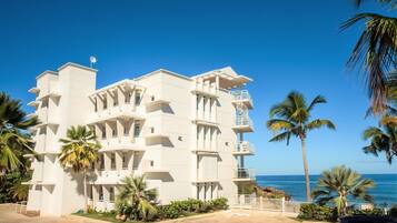 A multi-story hotel with balconies, located near a beach with palm trees.