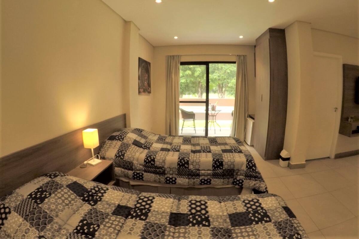 Een hotelkamer met twee bedden, een nachtkastje, een lamp, een deur naar een balkon en een raam met gordijnen.