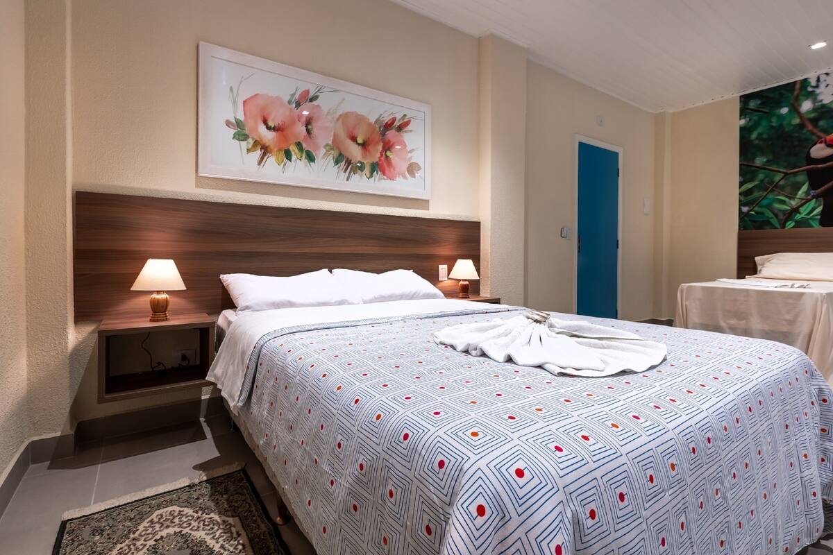 Hotelkamer met een groot bed, twee bedlampen, een houten hoofdeinde, een bloemschilderij, een blauwe deur en uitzicht op een boom door het raam.