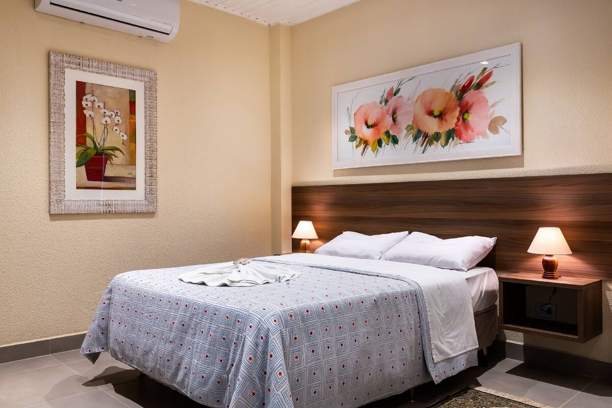 Een netjes opgemaakt bed met een bedsprei met patroon, twee nachtlampjes en een bloemschilderij in een lijst aan de muur.
