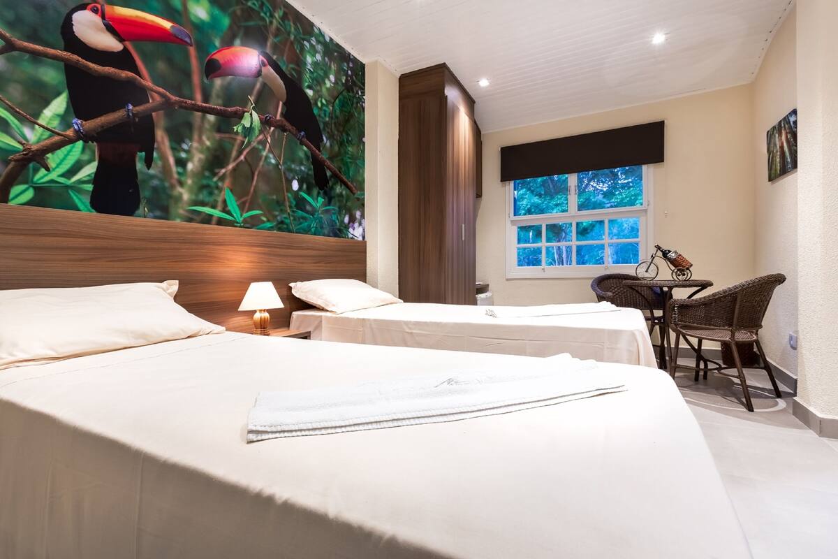 Hotelkamer met twee bedden, een grote muurschildering van toekans, een raam met uitzicht en een klein tafeltje met stoelen.