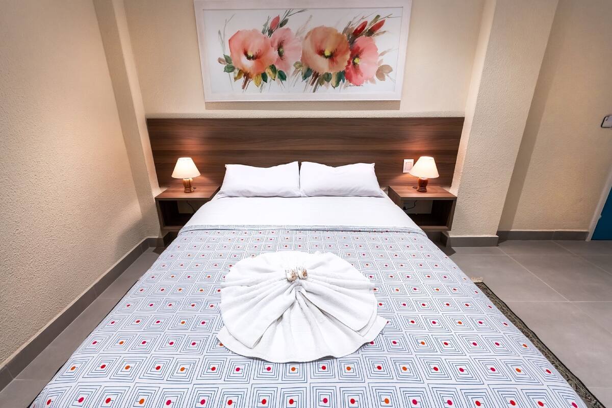 Een net opgemaakt bed met een beddengoed met patroon, twee bedlampen en een bloemschilderij aan de muur.