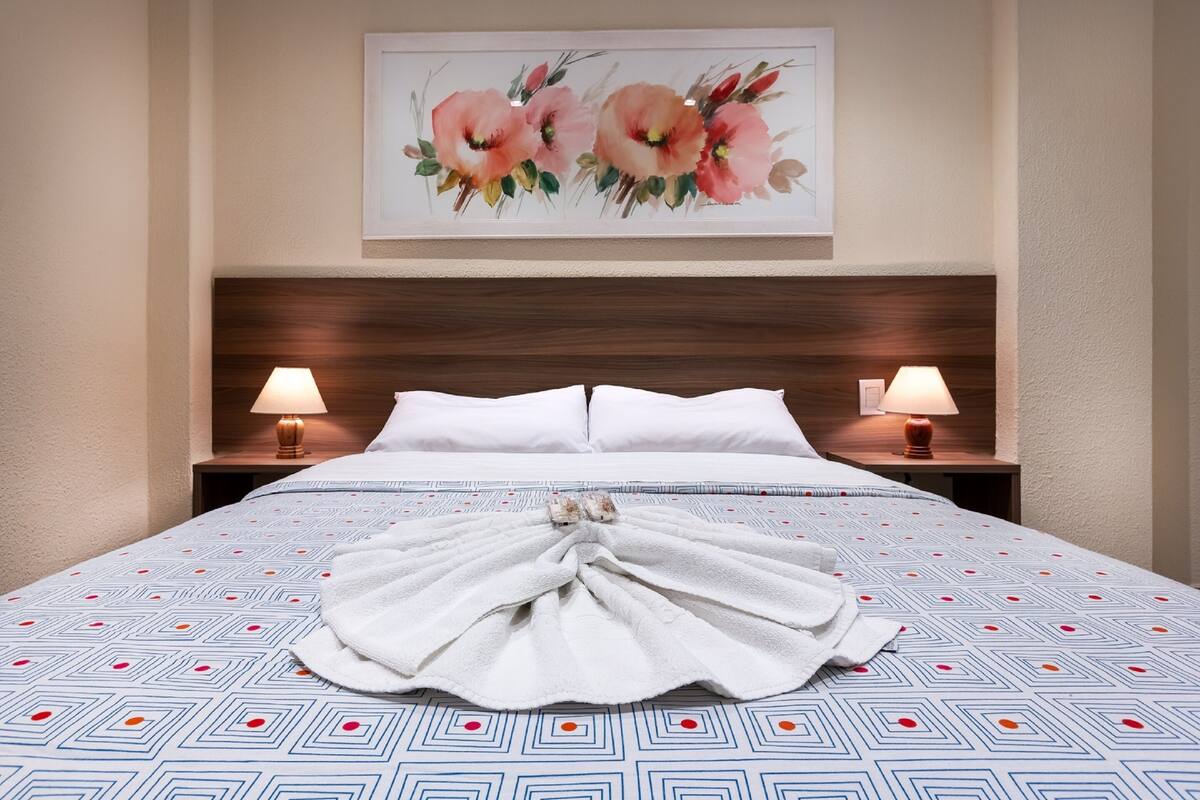 Een hotelkamer met een groot bed, twee bedlampen, een bloemistisch kunstwerk aan de muur en een handdoek op het bed.