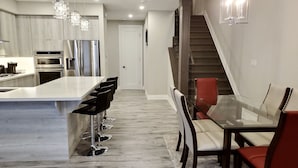 Una cocina moderna con electrodomésticos de acero inoxidable, una isla central con taburetes y un comedor con mesa y sillas de vidrio.