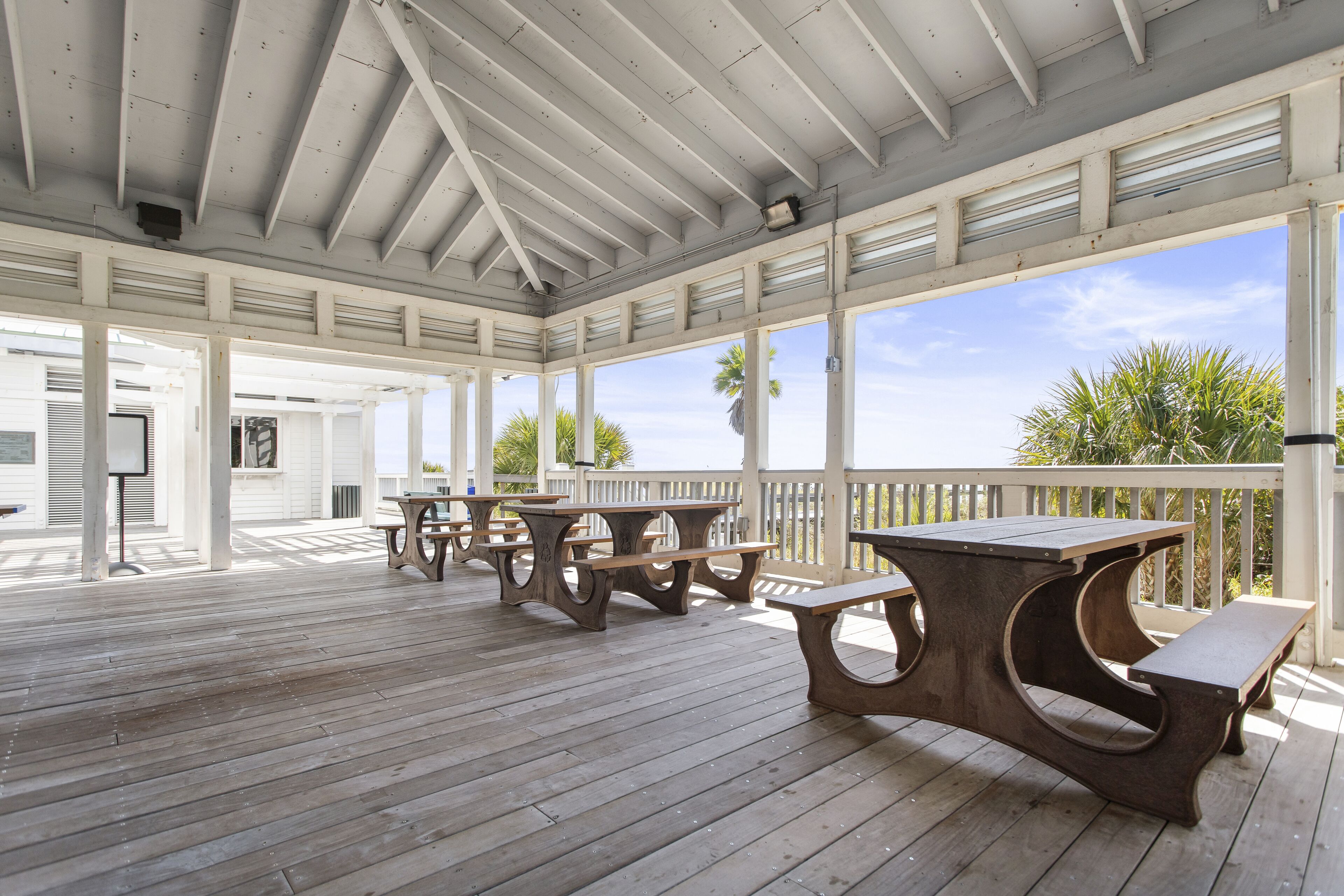 An outdoor covered patio with wooden tables and benches, white railings, and a view of palm trees.