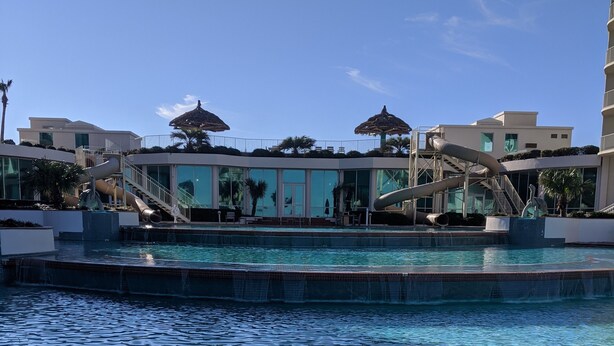 A swimming pool with a water slide, surrounded by modern buildings with large windows and outdoor seating areas.