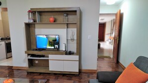 Ein Wohnzimmer mit einer Holzeinrichtungsanlage, einem Fernseher, einem Glastisch und einem Sofa mit orangefarbenem Kissen.
