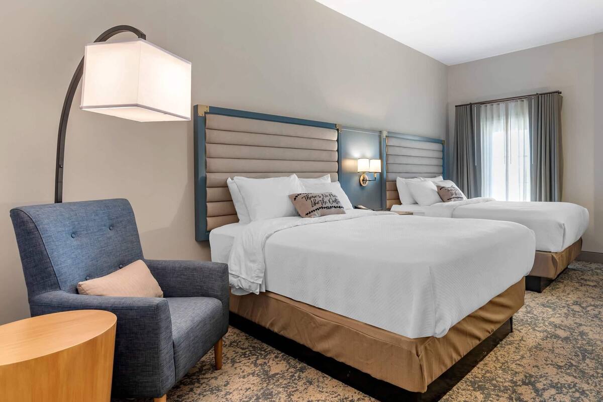 A hotel room with a large bed, a smaller bed, a chair, a lamp, and a window with curtains.