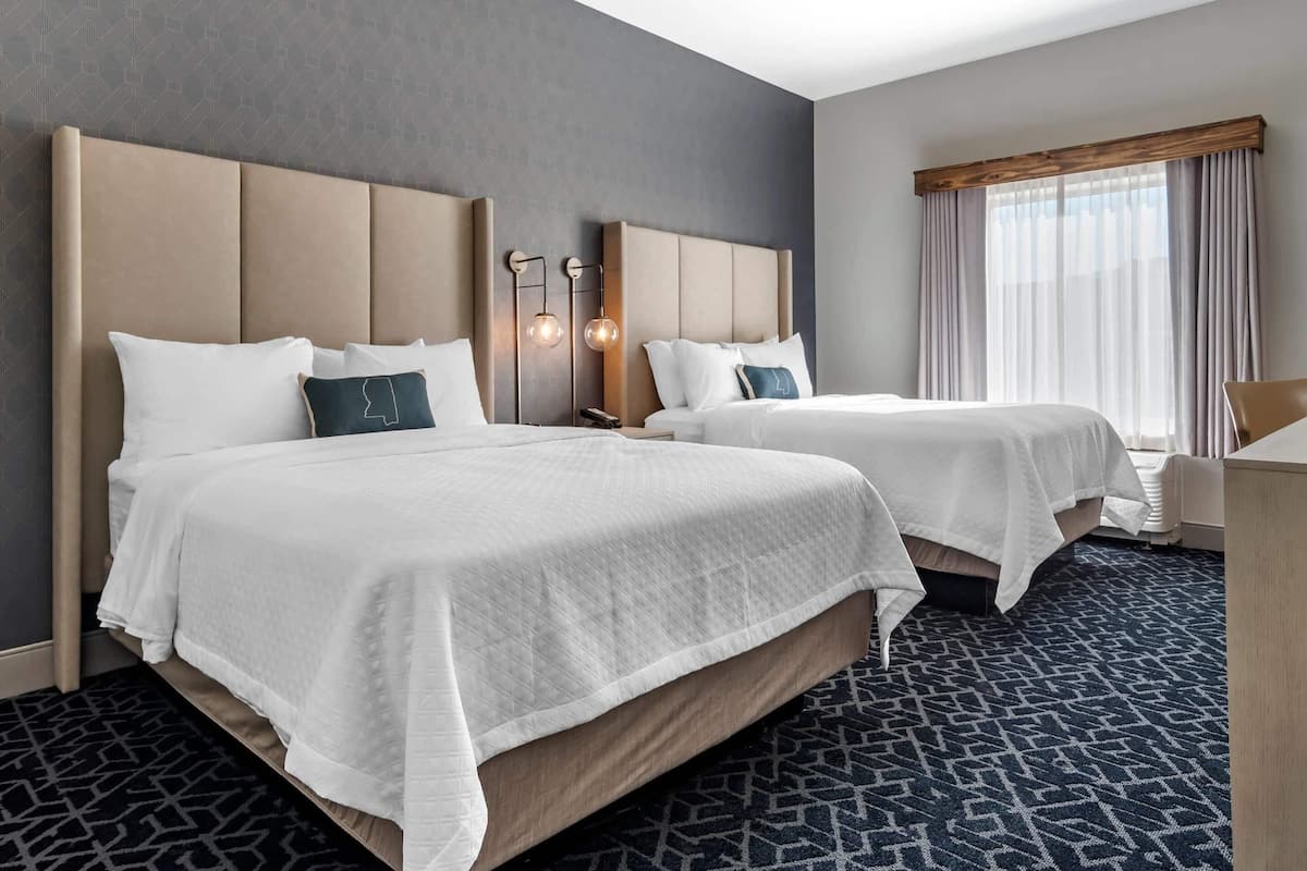 A hotel room with two beds, a large window with curtains, and a patterned carpet.