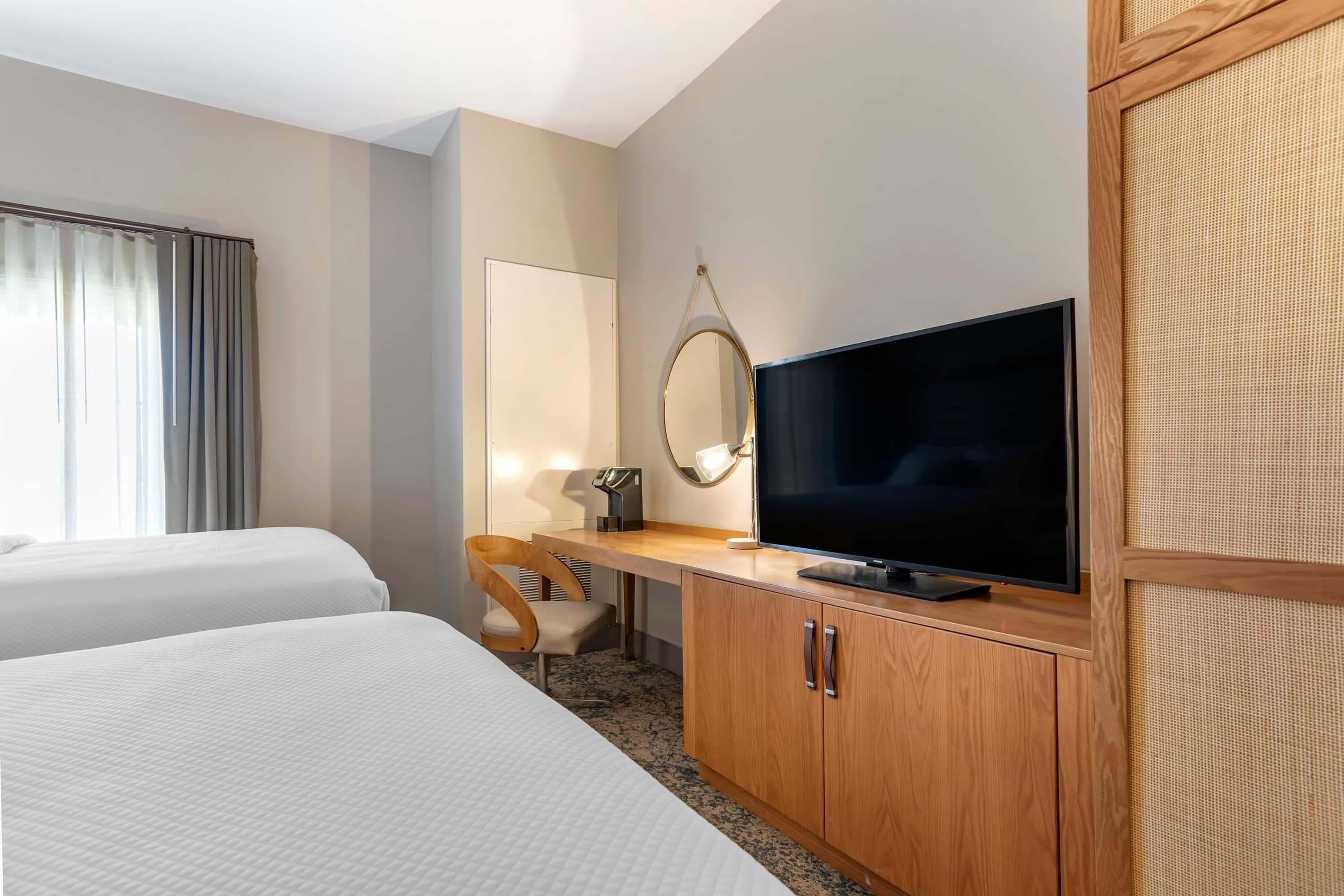 A hotel room with two beds, a TV, a desk with a chair, and a mirror.