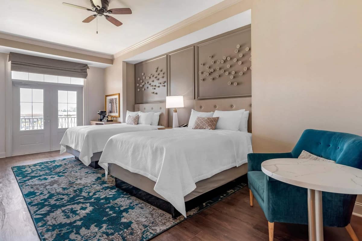 A hotel room with two beds, a blue armchair, a small round table, and a ceiling fan.