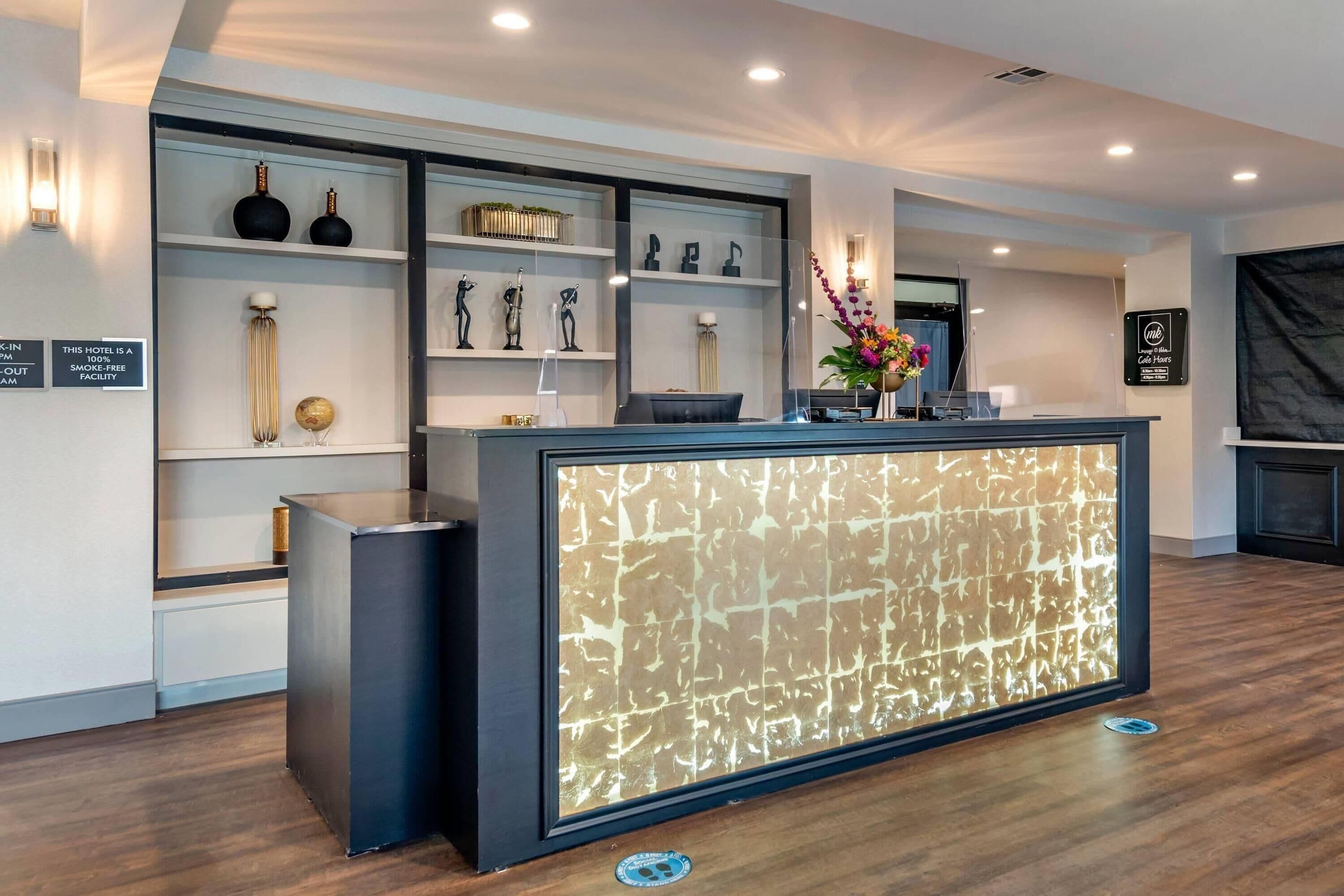 Un espace d’accueil d’hôtel au design moderne, comprenant un comptoir de réception agrémenté d’un panneau décoratif à motifs, des étagères ornées d’objets décoratifs et un panneau indiquant que l’hôtel est un établissement non-fumeur.