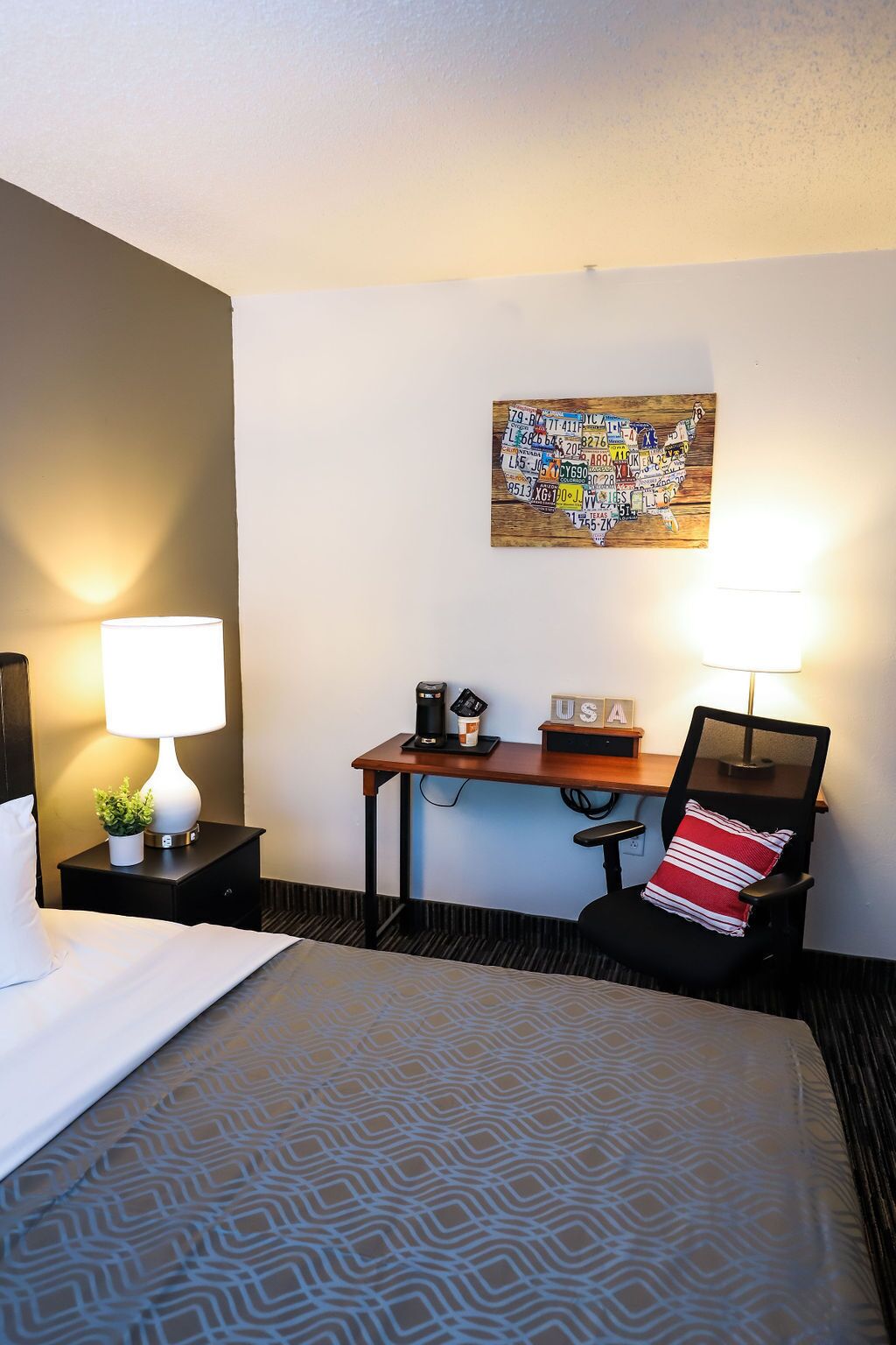 A hotel room with a bed, a desk, a chair, a map artwork on the wall, and a lamp on the nightstand.