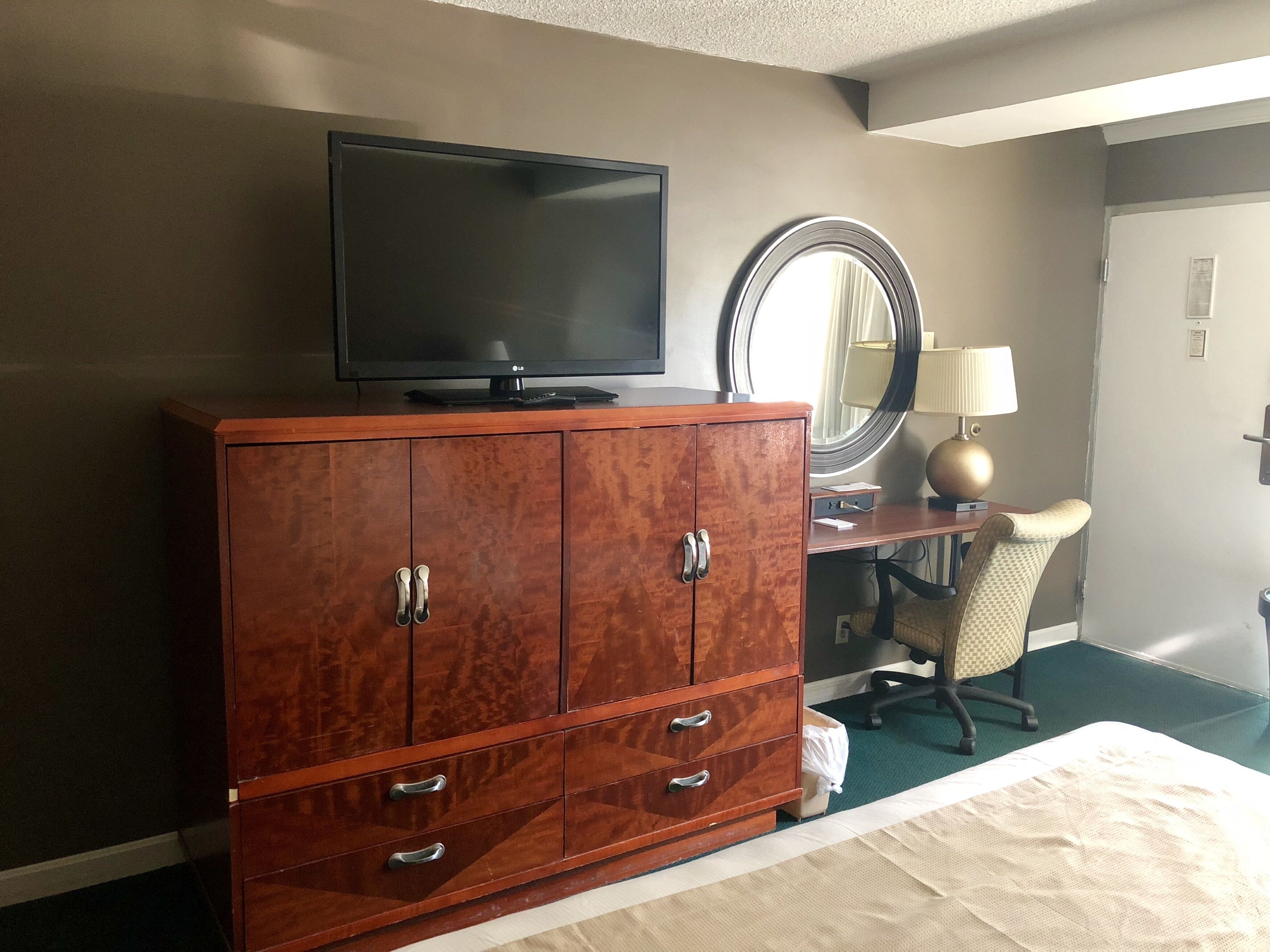 A hotel room with a wooden dresser, a flat-screen TV, a round mirror, a desk with a lamp, and a chair.