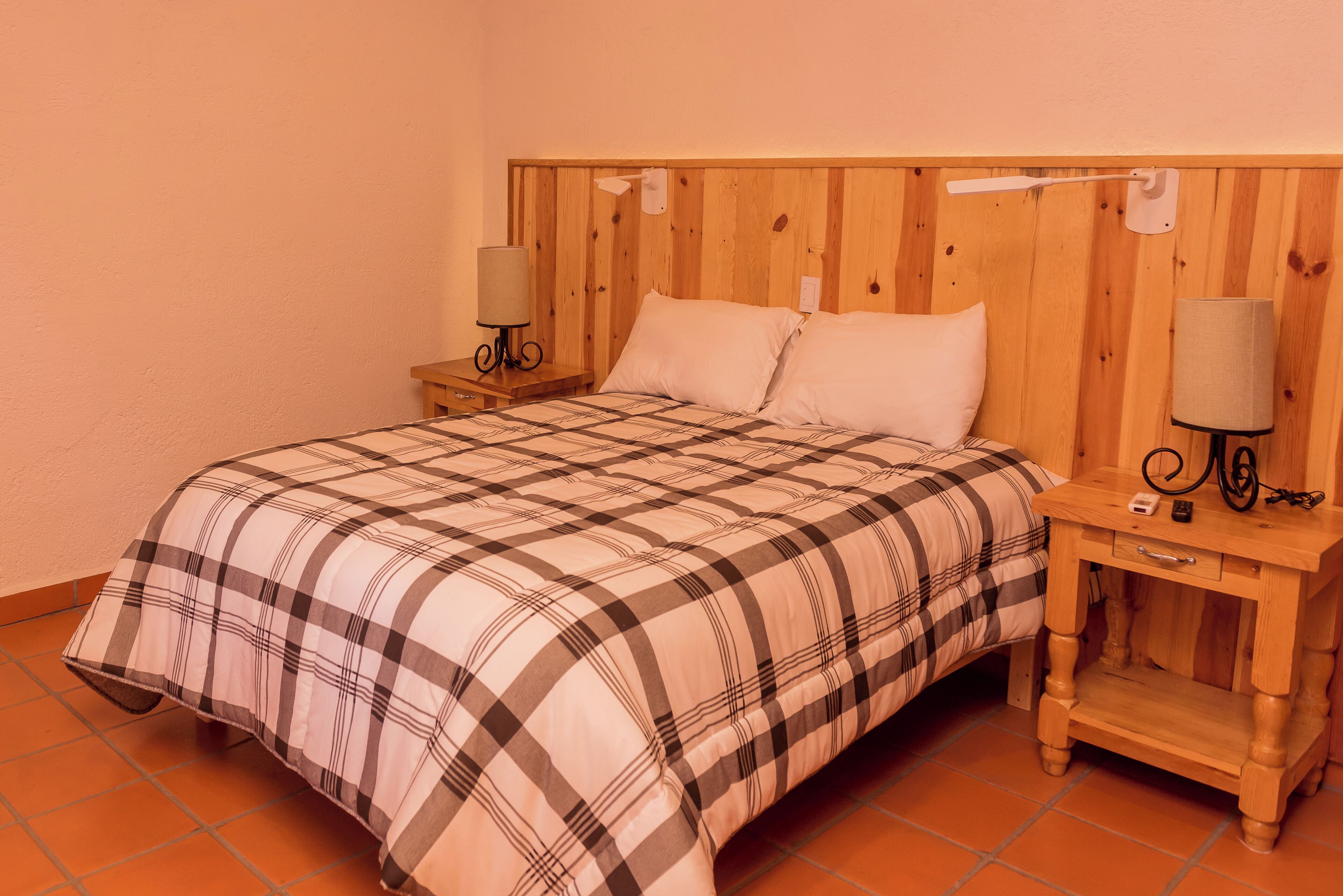 Un dormitorio con cabecera de madera, colcha a cuadros, dos mesitas de noche con lámparas y un pequeño aparador.