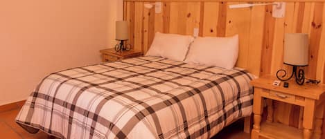 Un dormitorio con cabecera de madera, colcha a cuadros, dos mesitas de noche con lámparas y un pequeño aparador.