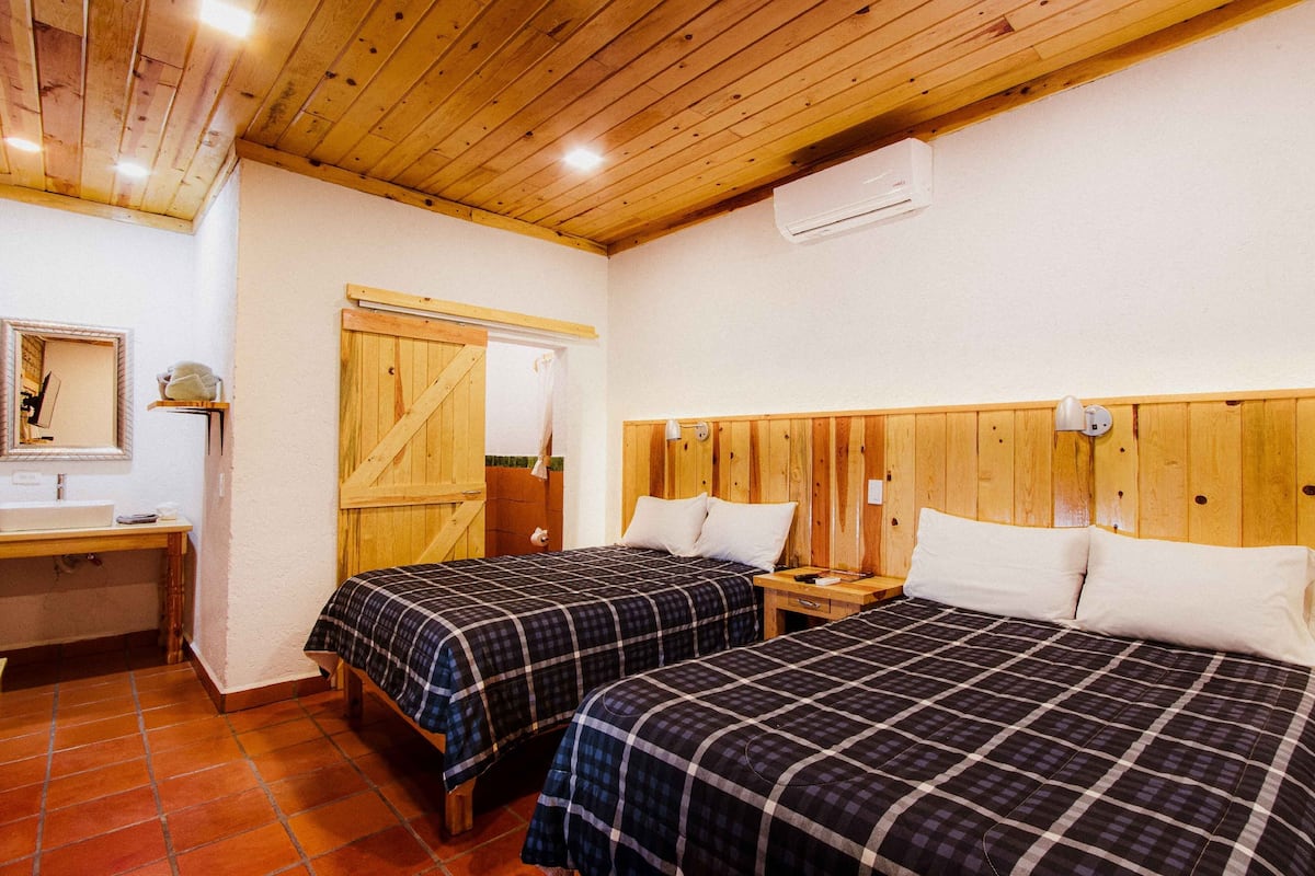 Een kamer met twee bedden, houten hoofdeinden, een klein nachtkastje, een spiegel en een raam met gordijn.